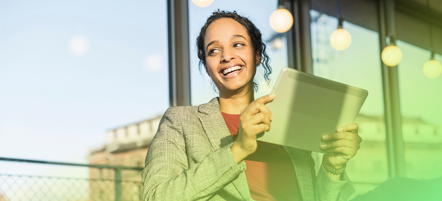 Lächelnde Frau im karierten Sakko sitzt vor großen Fenstern mit runden Hängelampen und hält ein Tablet in den Händen; das Bild ist mit einem grünen Farbverlauf überblendet.