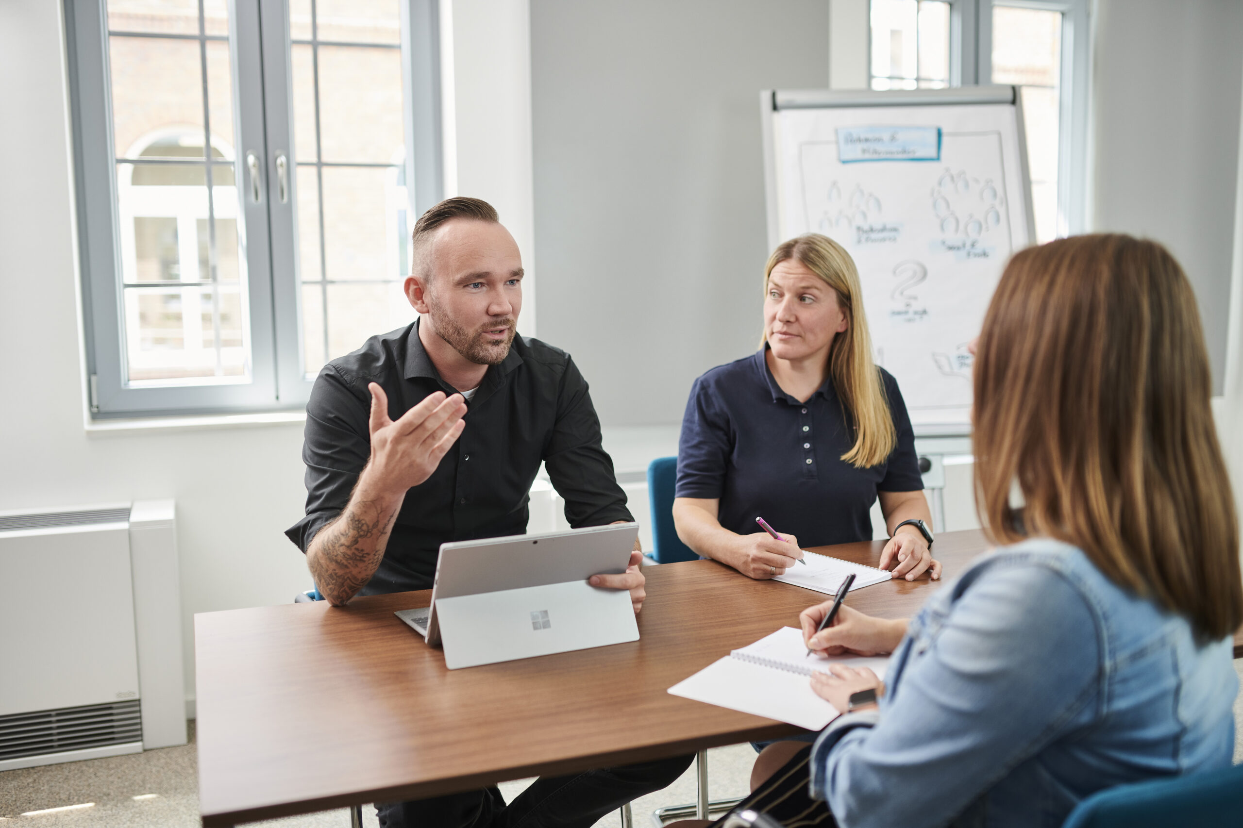 Ein Mann spricht in einer Besprechung, während zwei Frauen neben ihm Notizen machen.