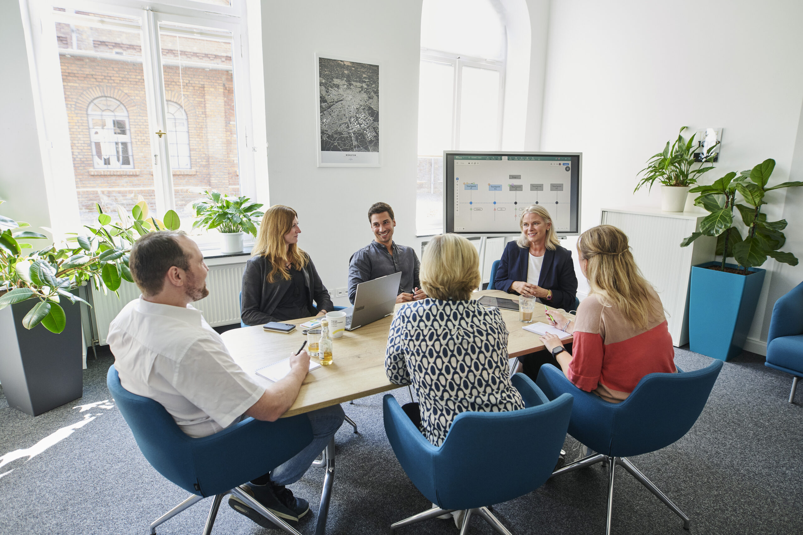 Sechs Personen sitzen in einem modernen Büro an einem Tisch und führen eine Besprechung. Auf einem Bildschirm im Hintergrund ist ein Diagramm zu sehen.