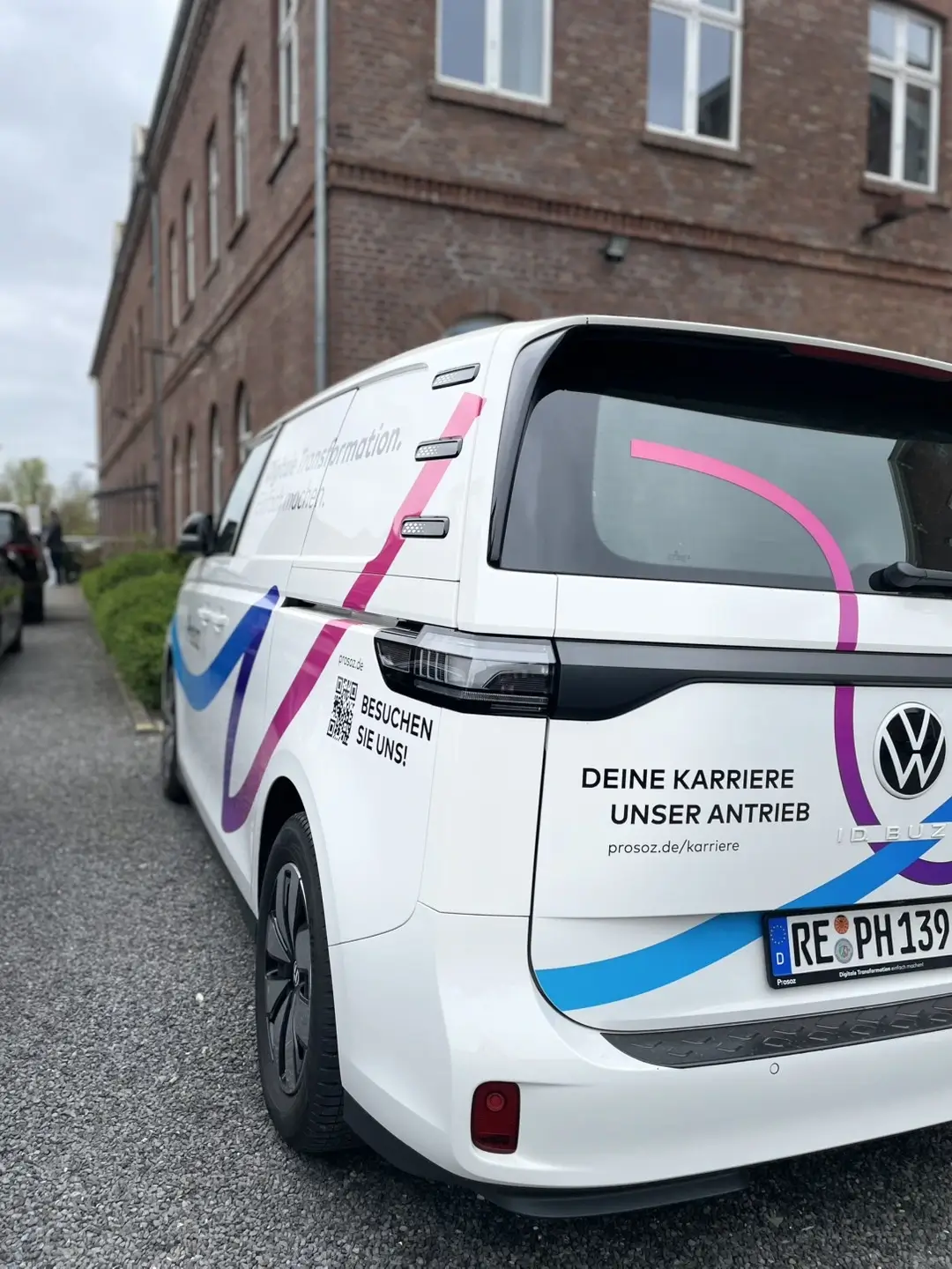 Weißer VW ID. Buzz mit farbigen Streifen und Werbeaufschrift „Deine Karriere – Unser Antrieb“ auf dem Heck. Das Fahrzeug steht vor einem Backsteingebäude auf einem Schotterparkplatz.