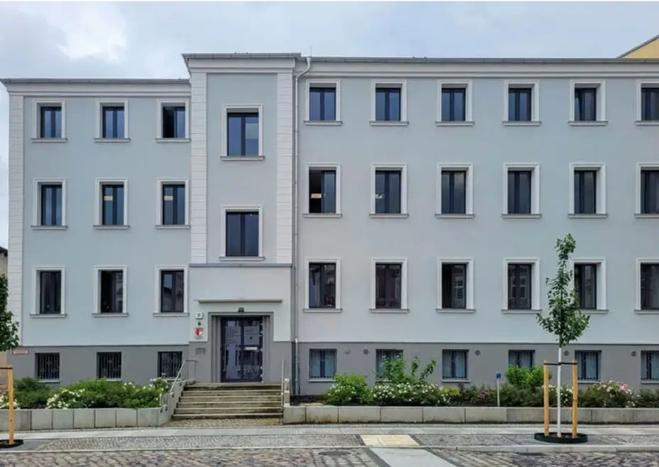 Fassade eines hellgrauen, vierstöckigen Gebäudes mit vielen gleichmäßig angeordneten Fenstern. Vor dem Eingang mit Treppe befinden sich Blumenbeete und junge Bäume.