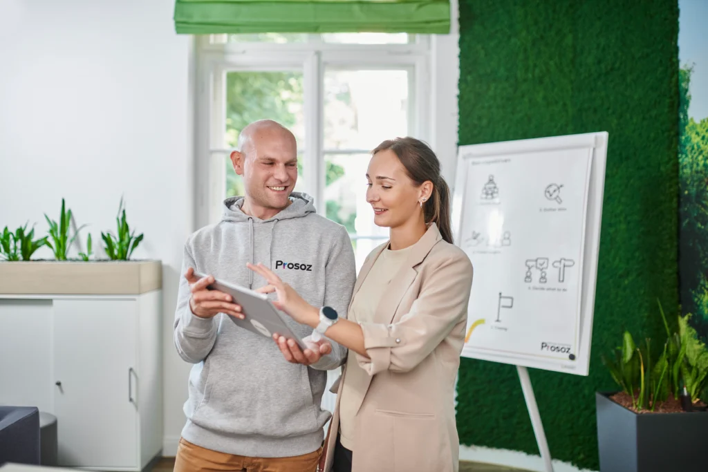 Digitale Verwaltung: Zwei Kolleg*innen betrachten gemeinsam ein Tablet; im Hintergrund Pflanzenwand und Flipchart.
