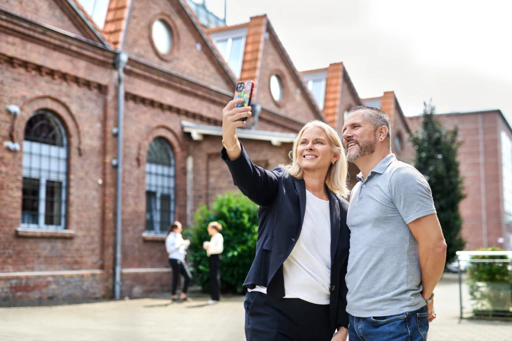 Zwei Kolleg*innen machen ein Selfie vor historischen Backsteinfassaden; weitere Personen im Hintergrund.