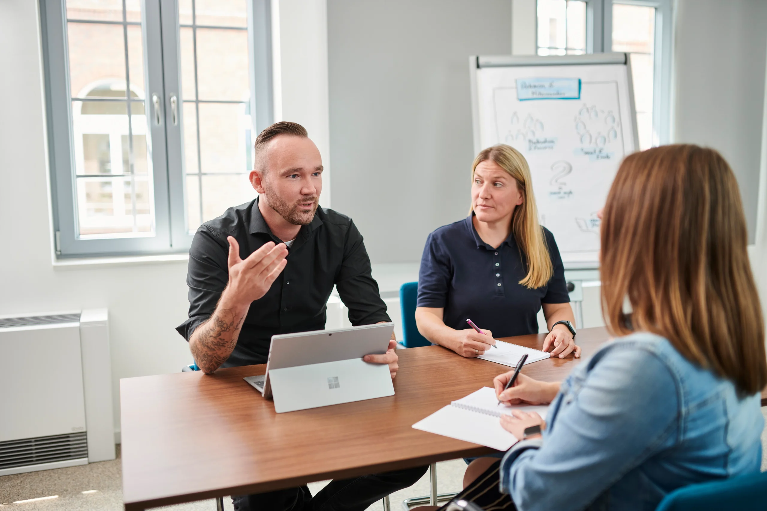 Drei Personen diskutieren am Tisch mit Notizblöcken und Tablet; Flipchart im Hintergrund