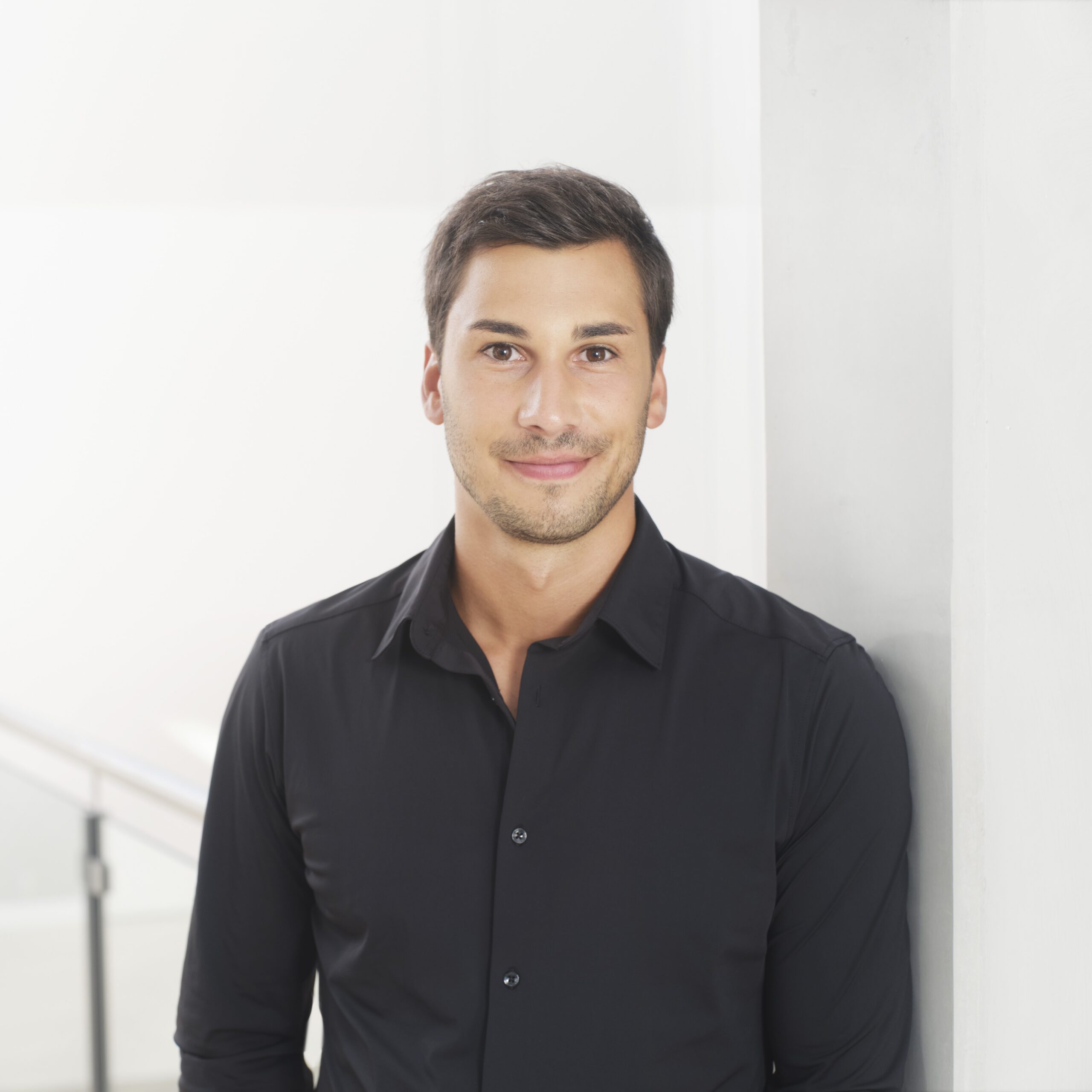 Business-Foto eines jungen Mannes im schwarzen Hemd, an eine helle Wand gelehnt